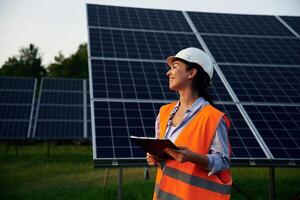 Black ,notepad, in hands. Female worker engineer in uniform is near solar panels photo