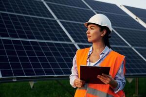 Black ,,notepad in hands. Female worker engineer in uniform is near solar panels photo