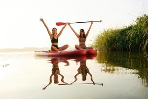 Hands up, smiling and looking,, forward. Women friends are on sup board on the lake together photo