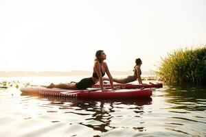 Asana, doing, yoga exercises. Women friends are on sup board on the lake together photo