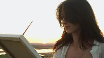 Young female artist with long dark curls is concentrating and meticulously running brush over canvas, creating work of art outdoors in rays of dawn sun. video