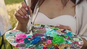 Portrait of young artist, holding bright multicolored palette in her hands, and pressing tip of brush against her lips in playful manner, coquettishly looking directly into camera. video