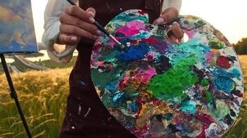 An artist, wearing an apron, carefully mixes oil paints on a colorful palette, standing in a field with a summer landscape as a backdrop. video