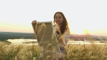 Attractive artist enjoys painting on canvas. She holds canvas in her hands and sits in middle of summer field with tall grass, with setting sun shining behind mountains as backdrop. video