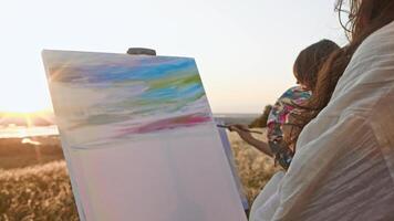 Pair of student artists paint landscape against the backdrop of the summer setting sun, in middle of hilly area near river, as part of their art school assignment. video