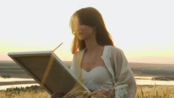 Art school student learns to paint landscapes while sitting on beautiful hill in field on summer evening, in rays of setting sun. video