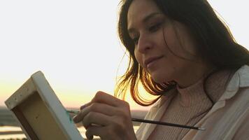 A left-handed street artist, sitting, carefully paints with a brush on canvas, using gouache, in the rays of the setting sun. Portrait. Slow motion video