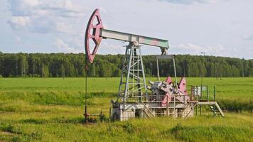 An old pumpjack, standing on the edge of a forest in the middle of a lush green meadow, is pumping oil from the depths below. video