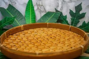 A beautifully crafted round tray of woven bamboo, showcasing intricate patterns and warm tones, set against a backdrop of lush green leaves, perfect for natural product presentations or rustic theme photo