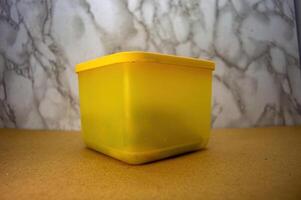 A plain yellow plastic container, subtly translucent, standing on a light brown surface against a blurred marble background, perfect for showcasing storage solutions or a minimalist aesthetic photo