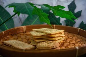 A close up view of golden biscuits and crackers neatly arranged in a rustic woven basket, framed by vibrant green foliage, perfect for a cozy snack or light refreshment photo