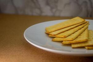 A close up of a stack of golden, rectangular crackers on a white plate, set against a brown surface and subtle textured background. Ideal for food content photo