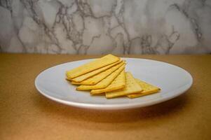A modest pile of golden, rectangular crackers arranged on a pristine white plate, set against a simple brown surface and textured wall. Ideal for food content photo