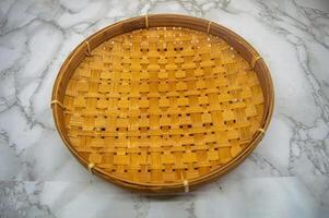 A traditional round woven bamboo tray or tampah, beautifully textured and resting on a white and gray marble surface, providing a versatile backdrop for natural or cultural themes photo