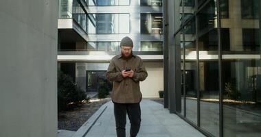 Young man with a beard, dressed in casual clothes and a knitted hat, is typing on a mobile phone while walking in the courtyard of a modern glass building. in 4k, red komodo photo
