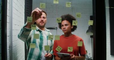 Caucasian man and an African American woman are planning, drawing a diagram of work on a glass board with stickers and using a tablet during a working day in the office. Brainstorm, business concept photo