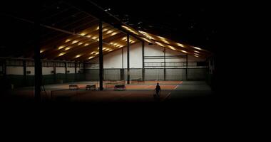 Large indoor sports hall with several tennis courts, lights are turned on and a man is seen walking towards the bench with a racket and a basket full of tennis balls in his hands. photo