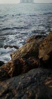 Sea waves hit large rocks on coast, foaming and splashing as they wash over the boulders. The hotel complex is visible on the peninsula in the distance, vertical . 4k , red komodo photo
