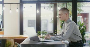 Young beautiful woman wearing glasses, dressed in casual clothes, working at table in a modern atmospheric cafe, making notes in a notebook and using a laptop photo