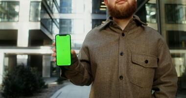 Mobile phone face forward in the hands of young man dressed in casual clothes, standing in the business center of the city. Copy space, green screen, chroma key, mockup. in 4k, red komodo photo
