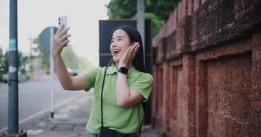 Handheld shot, Cheerful Asian woman happily takes a selfie while walking along a sidewalk next to a brick wall. video