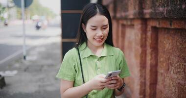 A young Asian woman is walking on the sidewalk and happily playing with her mobile phone. video