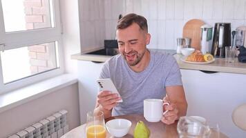 Man happily browsing messages or videos on his smartphone while having breakfast in the kitchen in the morning at home