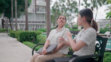 Relaxed Discussion Outdoors Two women are engrossed in conversation while sitting on a bench with trees and a swimming pool in the background, fostering a tranquil atmosphere. video