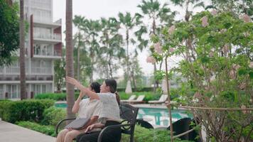 Two individuals sit on a bench and look up at buildings and palm trees, by the side of a pool. A scene filled with a sense of calm and thoughtful contemplation. video