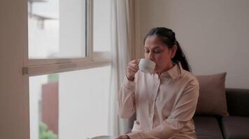 A middle aged woman is sipping a warm drink by the window where sunlight streams in and the soft light of the quiet atmosphere inside the house. video