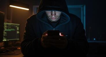 Anonymous hacker in dark hoodie using smartphone in dimly lit room with computer code. photo
