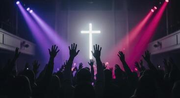 Silhouetted crowd with raised hands worships before a glowing cross under vibrant stage lights photo