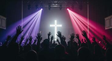 Crowd with raised hands at a worship event, illuminated by purple and pink spotlights and a glowing cross. photo