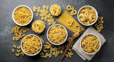 Overhead view of various dry pasta shapes in bowls and scattered on a dark surface photo
