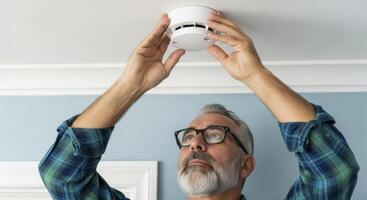 Mature man installing a white smoke detector on a ceiling for home safety photo