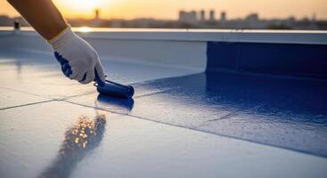 Worker applying blue liquid waterproofing coating with a roller on a rooftop surface at sunset photo