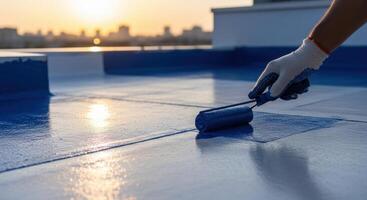 Worker applying blue liquid waterproofing coating with a roller on a building rooftop at sunset. photo