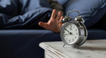 Hand reaching to silence a loud silver alarm clock on a nightstand in the morning. photo