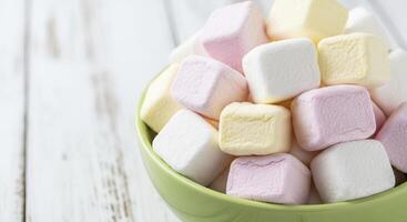 Close up of soft pastel cube marshmallows in a green bowl on a white wooden surface photo