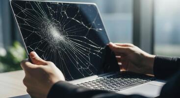 Close up of hands holding a dark gray laptop with a severely cracked screen photo