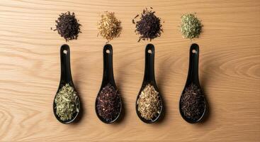 Four Black Ceramic Spoons Displaying Various Dried Tea Leaves on a Wooden Surface photo
