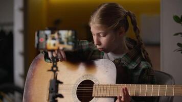 Talented Caucasian little girl guitarist singer playing guitar recording happy kid child blogger influencer filming online blog mobile phone camera tripod musical instrument song learning music video