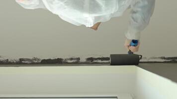 Woman in protective suit painting wall and ceiling with paint roller in black color during interior renovation video