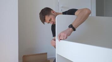 Man connecting parts of bed during home furniture assembly, surrounded by minimal tools and packaging. Handyman assemblies white bed frame on floor video