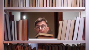 A man in glasses is peeking out from behind a book shelf video