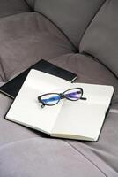 Pair of Stylish Black Glasses Rests on an Open Notebook Placed on Soft Gray Couch. photo