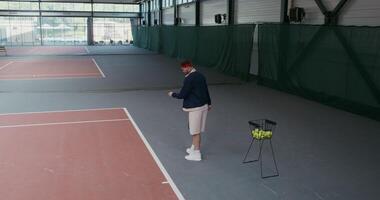 Man of European appearance takes a tennis ball from a basket full of balls and throws it over the tennis net, hitting the racket, playing in an empty indoor tennis court photo