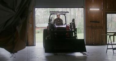Young man of European appearance with a beard drives a small agricultural vehicle with a bucket in front into a wooden garage, about to park after work on a farm on an autumn day photo