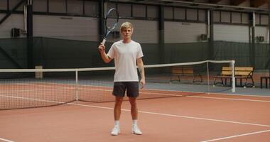 Young male tennis player of European appearance with a serious face throws a tennis racket behind his head, looking directly at the camera, standing on an indoor tennis court photo