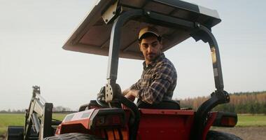 Young European farmer operates a farm tractor, while sitting in an open cab with canopy and turning back to control the evenness of the plowing strip photo